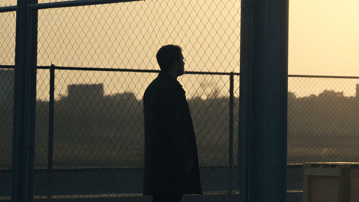 A lone figure watching from behind a chain-link fence at dusk.